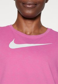 Růžové sportovní tričko s kulatým výstřihem, vyrobené z měkké tkaniny, s bílým logem Nike na přední straně. Jednoduchý design.