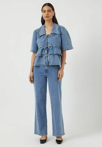 Sininen farkkutoppi lyhyillä puffihihnoilla, kerroksellisella peplum-muotoilulla, edessä solmittavalla kiinnityksellä ja yhteensopivat siniset levenevät farkut. Mustat korkokengät.