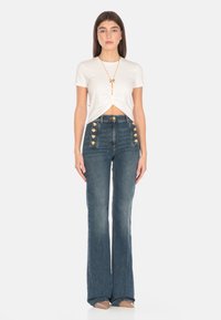 T-shirt bianco a maniche corte con dettaglio intrecciato sul davanti, abbinato a jeans flare in denim scuro a vita alta con dettagli con bottoni dorati.