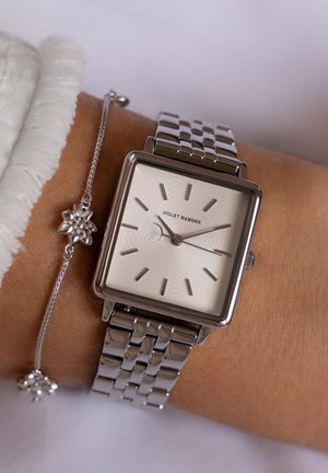 Montre carrée en acier inoxydable argenté avec un cadran blanc texturé, des index horaires argentés et un bracelet tressé. Le bracelet est orné d'un charm floral.