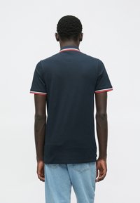 Να�υτικό πόλο μπλουζάκι με κόκκινη και λευκή ρίγα στον γιακά και τα μανίκια, φτιαγμένο από έναν λείο ύφασμα, εκτίθεται από πίσω. Χαλαρή εφαρμογή.