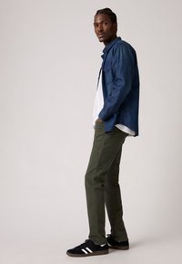 Camicia di jeans blu scuro con bottoni, t-shirt bianca sotto, pantaloni verde oliva e sneakers nere con strisce bianche. Stile casual.