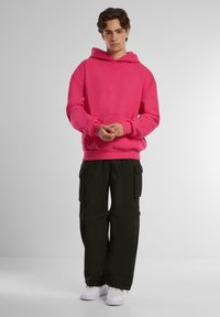 Sudadera con capucha rosa y bolsillo tipo canguro, combinada con pantalones cargo negros que tienen bolsillos laterales, y zapatillas blancas.