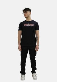 Zwart T-shirt met korte mouwen en het "CJ GAUDIJANS" logo, gedragen met zwarte pantalons en witte sneakers. Model staat in een neutrale pose.