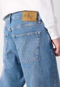 Lysblå denim jeans med en falmet look, der har baglommer og et brunt læder Calvin Klein-logo på livbåndet.