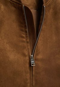 Veste en daim marron avec une fermeture éclair argentée, vue en gros plan. Le tissu a une texture douce et un design simple et épuré sans motifs.