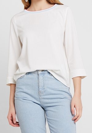 Vit långärmad topp med rund halsringning och kontrasterande kant, i kombination med ljusblå högmidjade jeans.