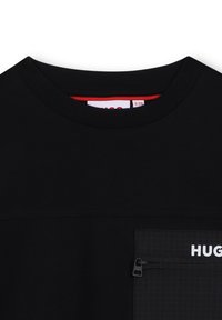 Camiseta negra de cuello redondo con un bolsillo en patrón de cuadrícula con cremallera en el pecho que presenta el texto parcial en blanco "HUGO".