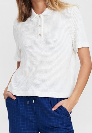 Femme portant un polo blanc texturé à manches courtes avec des boutons en perle et un pantalon à carreaux bleu, les mains dans les poches.