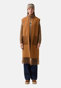 Brauner gestrickter Weste mit langem Schnitt, ausgestattet mit zwei gesteppten Taschen und fransigem Saum. Kombiniert mit einem beigen Rollkragenpullover und Jeans.