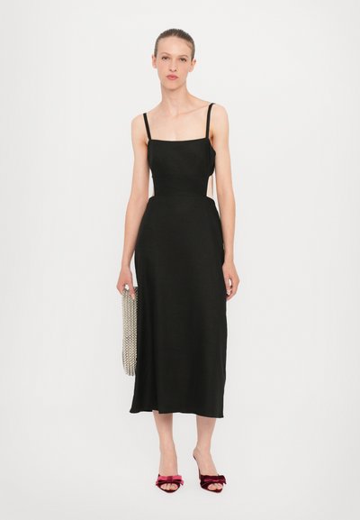 Zwarte jurk tot de knieën met dunne banden, een aangesloten taille en open zijkanten. Het model houdt een metallic kettingtas vast en draagt rode fluwelen muiltjes.