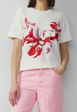 Vrouw draagt een wit shirt met korte mouwen met rode bloemen pailletten en roze broek, met één hand in de zak tegen een effen achtergrond.