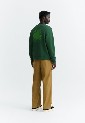 Personne debout de profil portant un sweat-shirt vert avec un motif circulaire en forme de rayons de soleil dans le dos et un pantalon large marron.