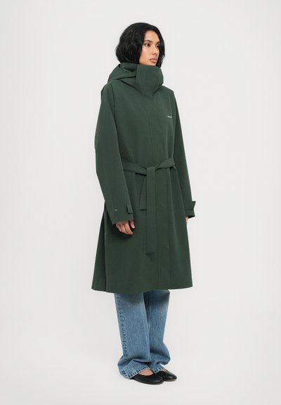Manteau vert à col montant, taille ceinturée et manches longues, associé à un jean bleu en denim ample et des chaussures noires. Texture lisse, design minimaliste.