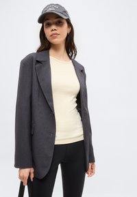 Młoda kobieta ubrana w szarą oversize'ową marynarkę, kremowy dopasowany top, czarne spodnie i szarą czapkę z daszkiem z napisem, stojąca na białym tle.