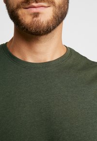 Tricou din bumbac verde măsliniu, cu guler rotund și mâneci scurte, având o textură netedă și un design simplu.