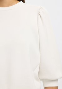 Hvit genser med korte puffermede ermer, myk tekstur og høy, rund hals. Minimalistisk design uten synlige mønstre eller detaljer.