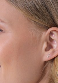 Silberne Ohrringe in Form eines dreißigenden Notenschlüssels mit minimalistischem Design, glatter Oberfläche und glänzendem Finish, die an einem menschlichen Ohr getragen werden.