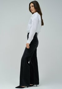 Mujer con cabello largo castaño, vistiendo una camisa blanca, jeans negros de pierna ancha y cintura alta, y tacones negros de punta, de pie contra un fondo liso.