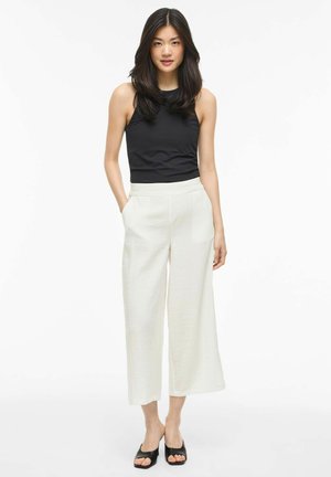 Femme aux longs cheveux foncés, portant un top noir sans manches, un pantalon blanc ample coupé court, et des talons ouverts noirs, debout les mains dans les poches.