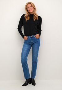 Svart ribbad långärmad topp med hög halsringning, matchad med blå raka jeans och svarta ankelstövlar, placerad mot en ljus bakgrund.