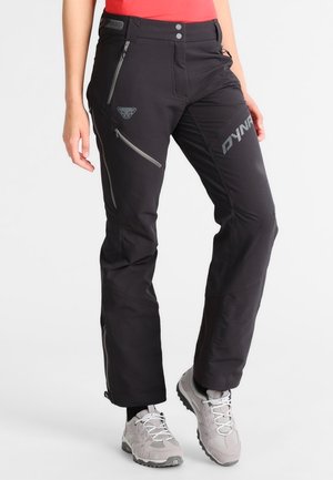Osoba ubrana w czarne spodnie outdoor z wieloma kieszeniami na zamek i szare buty do wędrówek, stojąca na jednolitym tle.