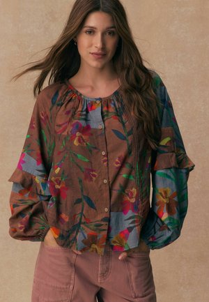 Ung kvinna med långt hår som bär en brun blus med blommönster och knappar samt rosa byxor, ståendes med händerna i fickorna mot en beige bakgrund.