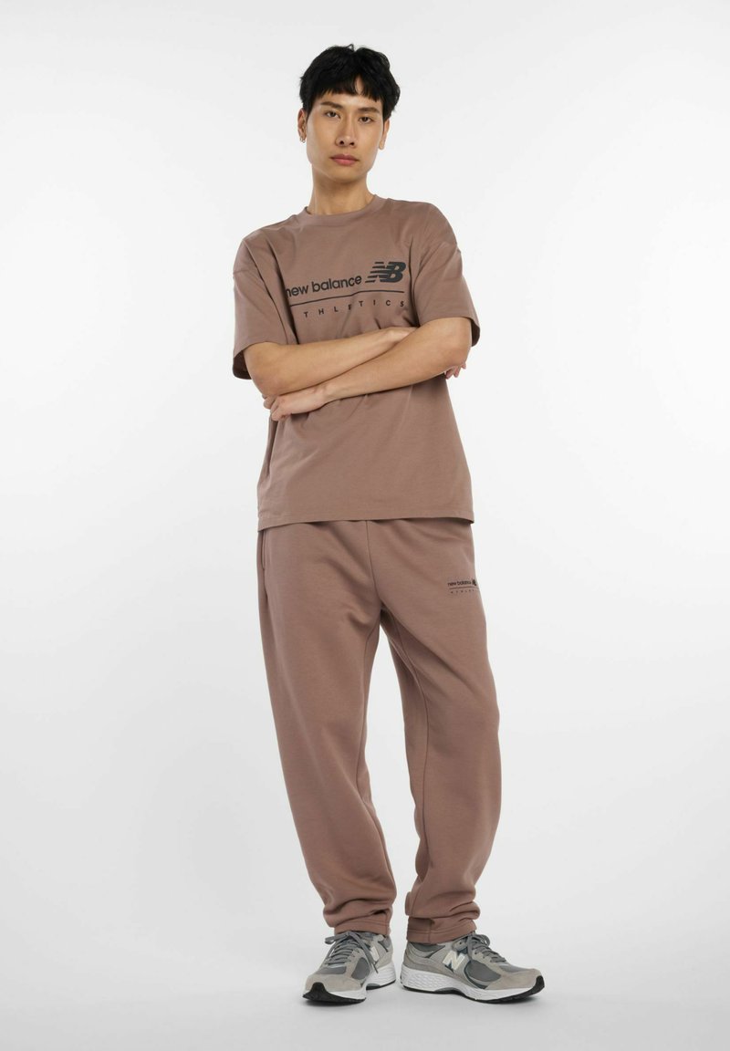 Camiseta de algodón marrón y pantalones de chándal a juego con el logo negro de "New Balance". Zapatillas grises con detalles blancos y suelas texturizadas.