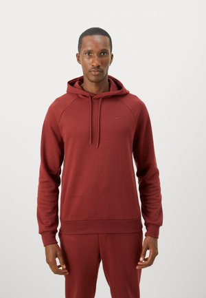 Man staat in een bijpassende bordeauxrode hoodie en joggingbroek tegen een effen lichte achtergrond, kijkt recht vooruit met een neutrale uitdrukking.