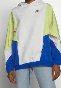 Felpa con cappuccio Nike caratterizzata da un corpo grigio con maniche a blocchi di colore verde, blu e bianco. Il materiale ha una texture morbida, coulisse regolabile all'orlo.