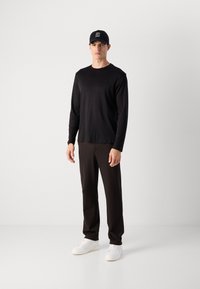 Mann iført svart langermet skjorte, svarte bukser, hvite joggesko og en svart Calvin Klein Jeans caps, som står mot en enkel hvit vegg.