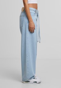 Osoba v modrých širokých džínách s denimovým páskem, bílé zkrácené tričko a bílé tenisky, stojící bokem na šedém pozadí.