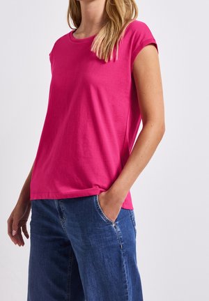 Kobieta w jasnoróżowej koszulce z krótkim rękawem, lekko włożonej do niebieskich dżinsów, z jedną ręką w kieszeni, na jednolitym tle.