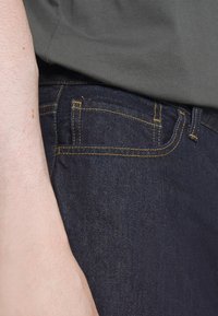 Ciemne dżinsy z żółtym przeszyciem, z przednią kieszenią i gładką fakturą. Szara koszula częściowo zasłania pas biodrowy.