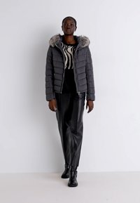 Giubbotto puffer grigio con cappuccio in ecopelliccia, abbinato a un maglione nero con un motivo bianco e pantaloni in pelle nera. Il modello indossa stivali neri alla caviglia.