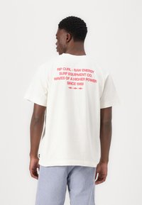Cremfarbene Baumwoll-T-Shirt mit rotem Text auf der Rückseite, der die Marke und den Slogan angibt. Das Modell trägt eine blau-weiß gestreifte Hose.