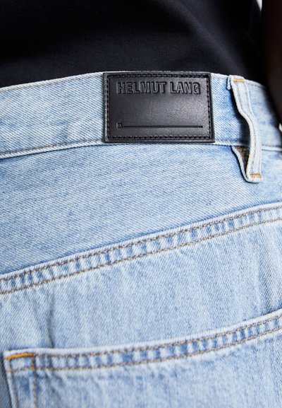 Ανοιχτού μπλε τζιν παντελόνι με μαύρη δερμάτινη ετικέτα που φέρει ανάγλυφο το "HELMUT LANG" στη μέση και ορατές ραφές στις τσέπες.