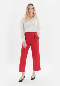 Röda högmidjade byxor med sidofickor och svarta knappdetaljer, tillsammans med en vit långärmad blus och svarta spetsiga skor.