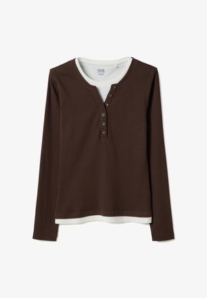 Chemise henley marron à manches longues superposée à un haut à col rond blanc avec des boutons visibles sur la patte de la chemise marron.