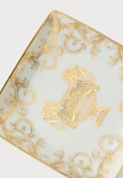 Assiette carrée en porcelaine blanche avec des ornements dorés en forme de volutes et deux chérubins tenant un tissu drapé au centre.