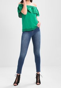 Zelený top bez ramínek s krajkovým lemem, v kombinaci s těsnými modrými džíny a černými šněrovacími podpatky. Jednoduché pozadí.
