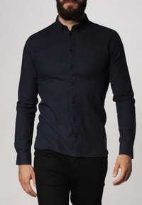 Navyblauwe overhemd met lange mouwen, knoopsluiting en kraag, voorzien van een borstzak en donkere knopen. Soepele textuur, gecombineerd met zwarte broek.