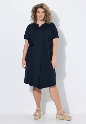 Mujer con cabello rizado, vestida con un vestido azul marino hasta la rodilla con mangas cortas y sandalias plataforma beige, de pie sobre un fondo blanco.