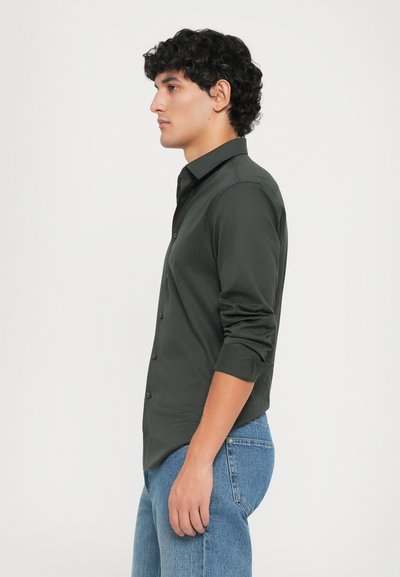 Cămașă verde închis cu mâneci lungi, cu un inserție butonată și guler clasic, purtată cu blugi de un albastru deschis. Textură fină, design ajustat.