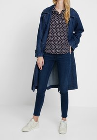 Cappotto lungo in denim, blusa blu marine a fantasia, jeans scuri e sneakers bianche. Le caratteristiche includono dettagli con bottoni e un colletto sulla blusa.