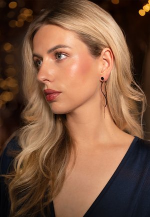 Orecchini pendenti neri e dorati con un design ondulato, dotati di un bottone circolare e filamenti allungati. Il modello ha lunghi capelli biondi e indossa un top scuro.