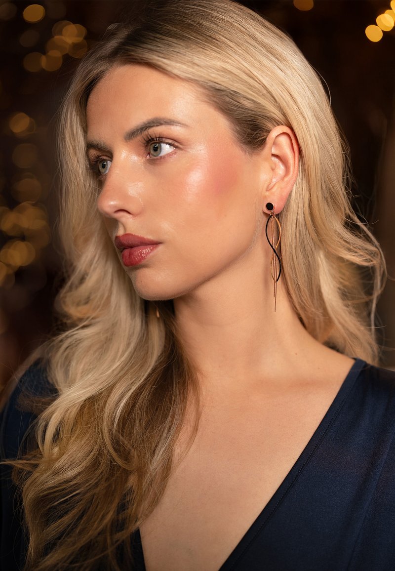 Black and gold dangle earrings featuring a wavy design, with a circular stud and elongated strands. Model has long blonde hair and wears a dark top.