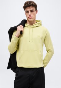 Νέος άνδρας που φοράει ανοιχτό κίτρινο φούτερ με μπροστινή τσέπη, κρατώντας ένα μαύρο μπουφάν στον ώμο του, στέκεται μπροστά σε μία απλή φόντο.