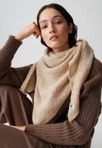 Beige, zacht getextureerde sjaal, draped om de hals, met een subtiel patroon en een klein merketiket, gedragen bij een bruine ribbeltrui.
