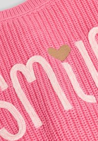 Pink knitted fabric features the word "Smile" in white, with a small beige heart accent. The texture is ribbed and soft.